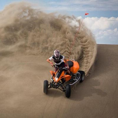 A person wearing a helmet and protective gear rides an orange ATV at the Killpecker Sand Dunes in 怀俄明. The ATV tires kick up sand and create a wavy wall behind it.